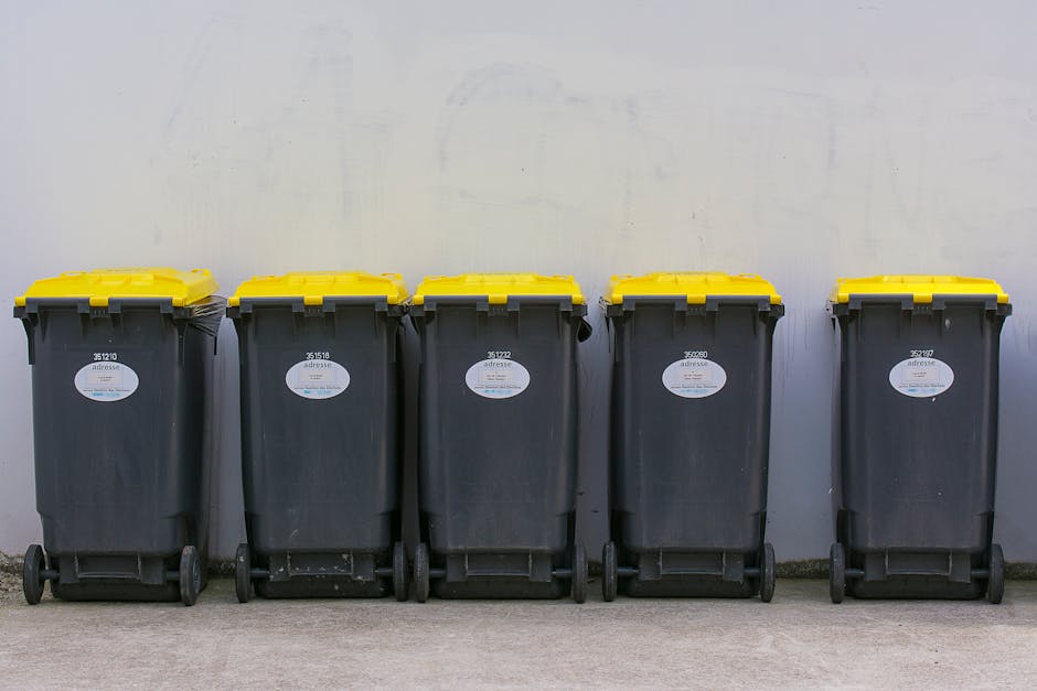 Two large black plastic bags filled with waste material are placed on a concrete sidewalk next to a low curb in an outdoor urban setting. The bags are crumpled and appear to contain household or yard