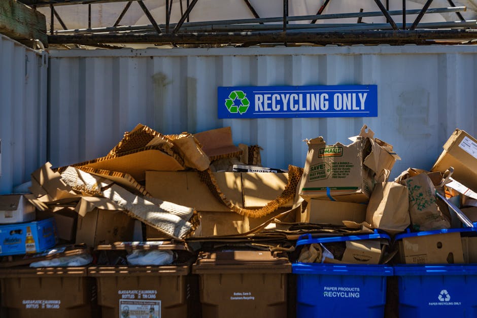 A collection of discarded cardboard boxes and paper packaging materials piled on top of reusable recycling bins, with some flattened and others crumpled, located outdoors against a corrugated metal wa
