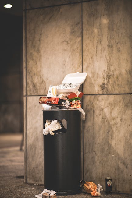 A collection of discarded cardboard boxes and paper packaging materials piled on top of reusable recycling bins, with some flattened and others crumpled, located outdoors against a corrugated metal wa