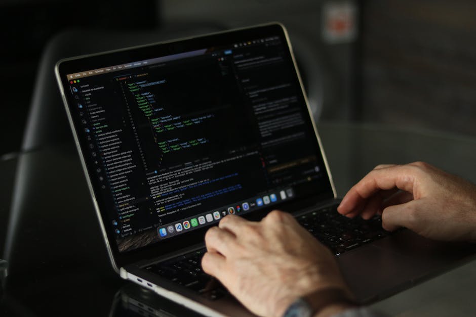 A person using a laptop computer with the screen displaying lines of code, with their hands positioned on the keyboard. The laptop is placed on a dark, glossy surface, possibly a desk, with a blurred 
